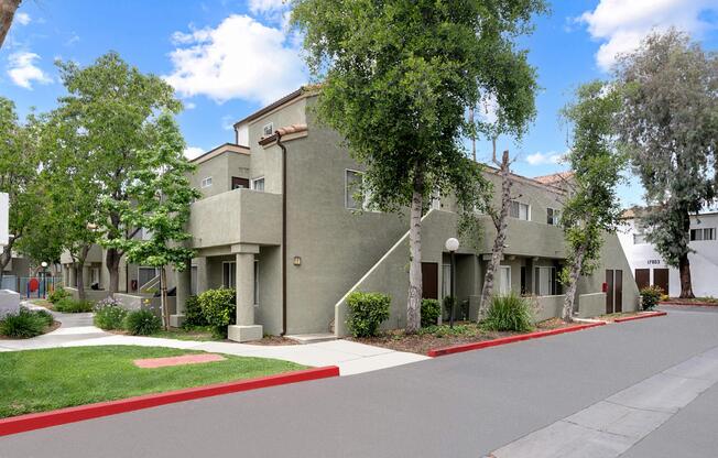 A peaceful residential area featuring modern two-story apartment buildings surrounded by trees and greenery. The pathway is bordered by well-maintained landscaping, and the street is clean with a red curb. Blue skies and fluffy clouds create a serene atmosphere.