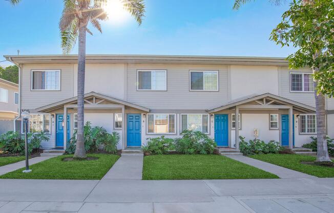 A row of modern townhouses with beige siding and blue front doors, flanked by palm trees. The grassy front yard is well-maintained, with shrubs and plants. Bright sunlight shines from the left, creating a warm, inviting atmosphere.