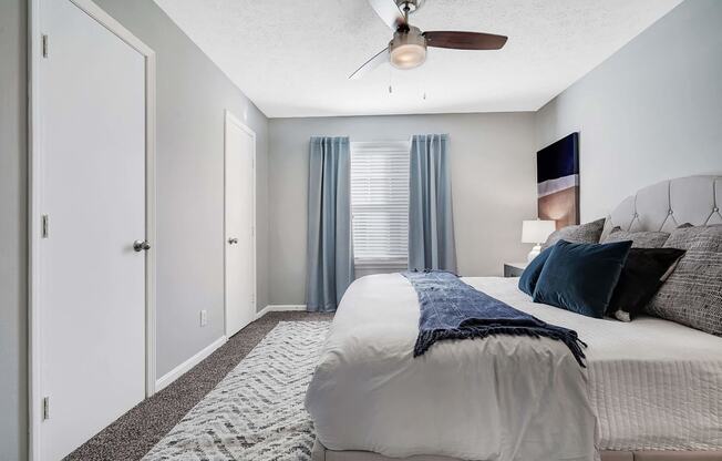 Apartments In Louisville, KY With In-Unit Washer And Dryer - Enclave At Breckenridge - Spacious Bedroom With Carpet Flooring, Large Bed, Nightstand with Lamp, Ceiling Fan, And Window.