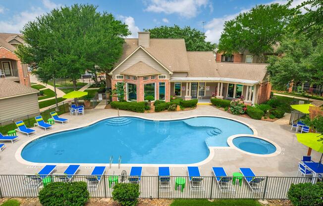 Aerial view of a residential pool area featuring a large, curvy swimming pool surrounded by lounge chairs and green umbrellas. The pool is bordered by well-maintained gardens and a modern building with large windows. Lush trees and shrubs enhance the inviting atmosphere of the outdoor space.