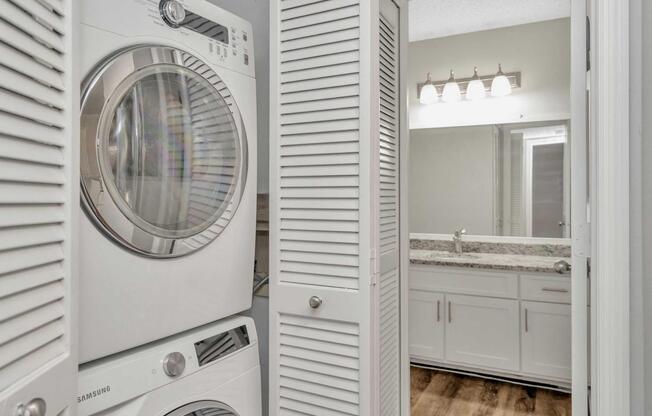 A compact laundry area featuring a stacked washing machine and dryer. In the background, a well-lit bathroom with a granite countertop and white cabinetry is visible, along with a mirror and light fixtures. The space is organized and has a modern design.