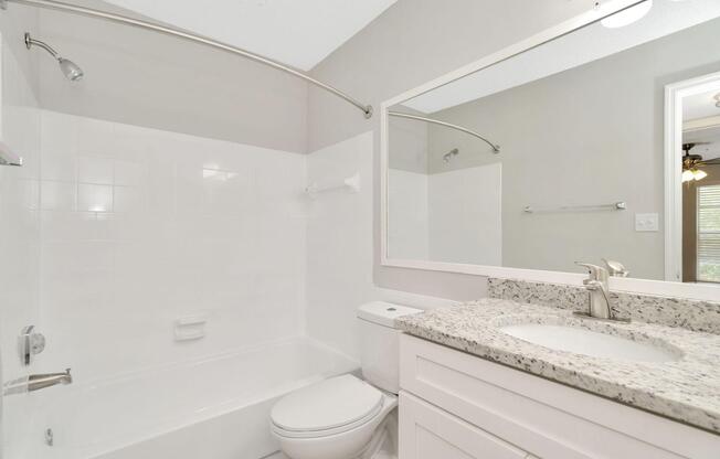 A clean, modern bathroom featuring a white tiled bathtub with a chrome shower rod, a sleek toilet, and a vanity with a granite countertop and a large mirror. The walls are painted in a light color, and thereâs natural light coming from a nearby window.