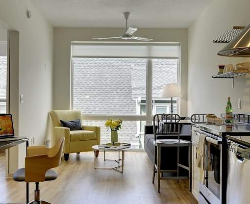 Living space with desk nook, yellow accent chair, grey couch and glimpse of kitchen in the Flat E floor plan at Coze Flats