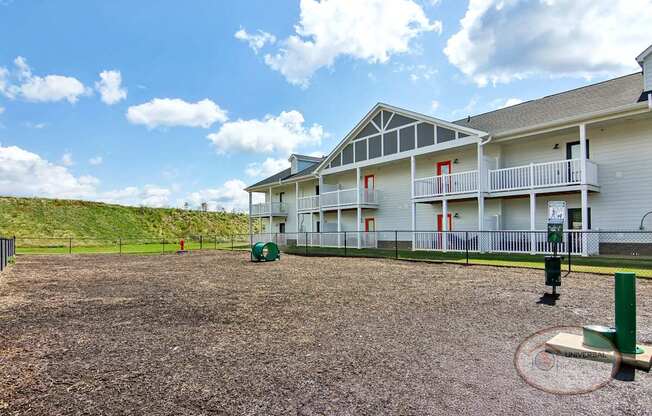 A fenced in dog park with a water fountain and obstacles for your dog with an apartment building in the background.