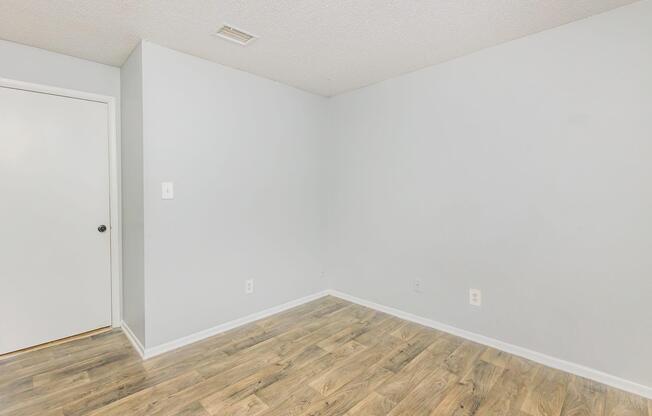 Empty room with light gray walls and a wood-like laminate floor. A single white door is visible on the left. The room is well-lit and spacious, featuring minimal decor and no furniture, creating an open and clean appearance.