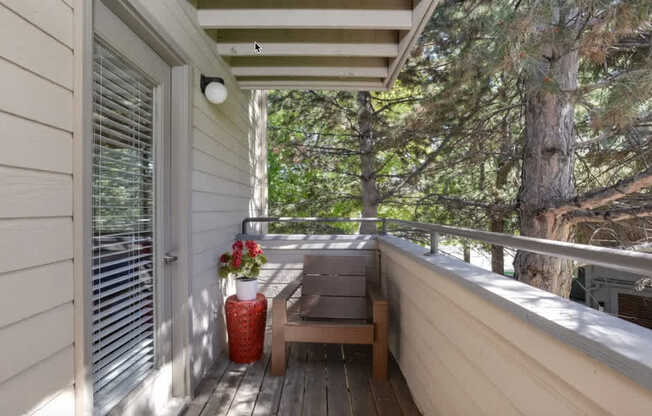 A wooden bench sits on a porch with a red vase and a tree in the background.