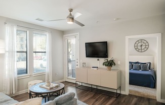 A living room with a grey couch, a round table, a television, and a bed with a blue comforter.