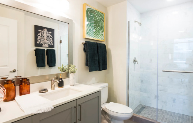 Bathroom with a glass-walled walk-in shower with floor-to-ceiling marble tile, a white quartz countertop with gray cabinets, and a large framed mirror.