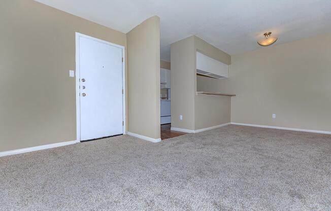 An empty living room with light beige walls and a carpeted floor. A white door leads outside on the left, with a small kitchen area visible in the background. The room has a warm light fixture on the ceiling and minimal furnishings, creating a spacious and inviting atmosphere.