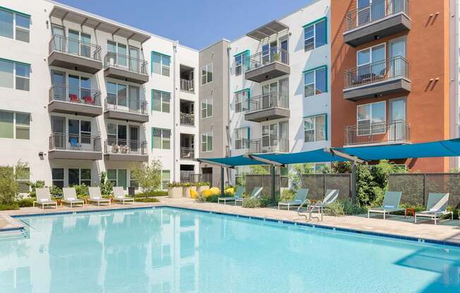 A swimming pool is surrounded by lounge chairs and umbrellas in front of apartment buildings.