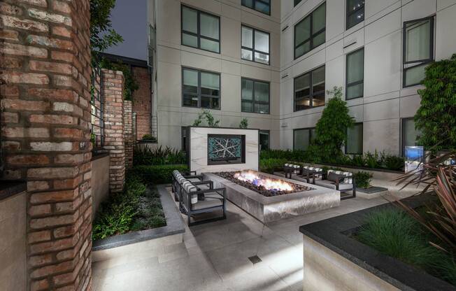 A landscaped stone courtyard with a large rectangular fire pit, 3 armchairs on each side, and an HDTV mounted on a freestanding stone wall.