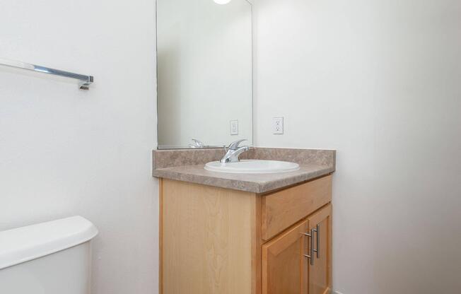 A clean and simple bathroom featuring a wooden vanity with a sink and a mirror above it. The countertop is light-colored, and the walls are painted white. A toilet is positioned to the left of the vanity, creating a functional and minimalist space.