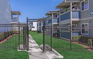 A landscaped area with a gated entrance leading to a pathway. On either side, there are two multi-story apartment buildings featuring balconies and blue accents. Lush green grass surrounds the pathway, creating a welcoming outdoor space. Clear blue skies are visible above.
