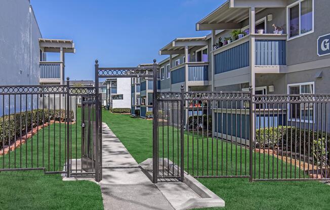 A landscaped area with a gated entrance leading to a pathway. On either side, there are two multi-story apartment buildings featuring balconies and blue accents. Lush green grass surrounds the pathway, creating a welcoming outdoor space. Clear blue skies are visible above.