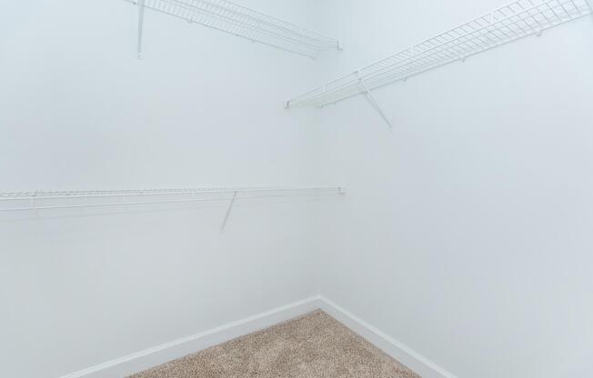 An empty closet with white walls and carpeted floor, featuring three wire shelving units mounted on the walls, offering storage space. The overall look is clean and minimalist, with no items present.