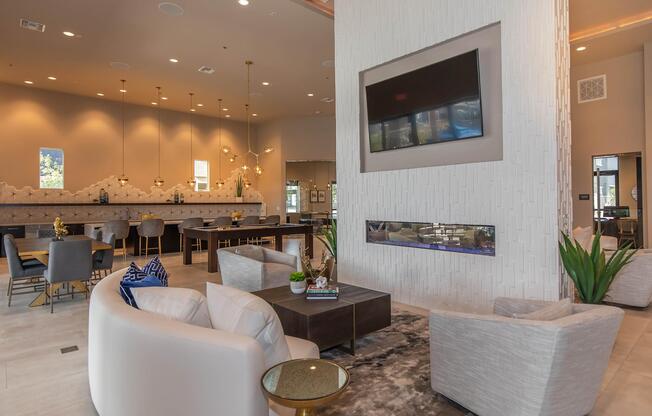 A modern, stylish lobby featuring two white armchairs and a coffee table in front of a contemporary fireplace. The walls are adorned with elegant decorations, and a large television is mounted above. Soft lighting creates a warm atmosphere, with dining tables and chairs visible in the background.