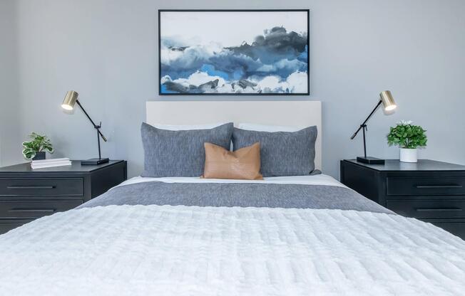 A modern bedroom featuring a neatly made bed with gray and white bedding, two bedside tables with table lamps, and small plants. Above the bed, there is a large abstract painting in shades of blue and gray, adding a touch of color to the neutral room.