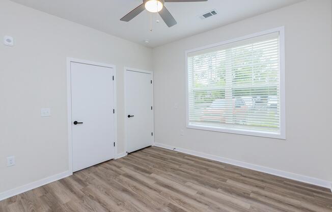A clean, empty room featuring light-colored walls, a ceiling fan, and two closed white doors. A large window with blinds allows natural light, illuminating the wooden floor. The space is minimalist and ready for furniture or decoration.
