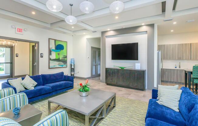 A modern living area featuring blue sofas, a coffee table, and a decorative plant. The room has stylish lighting fixtures and a large flat-screen TV on the wall. Light-colored walls and a patterned carpet create a welcoming atmosphere. A kitchenette is visible in the background.