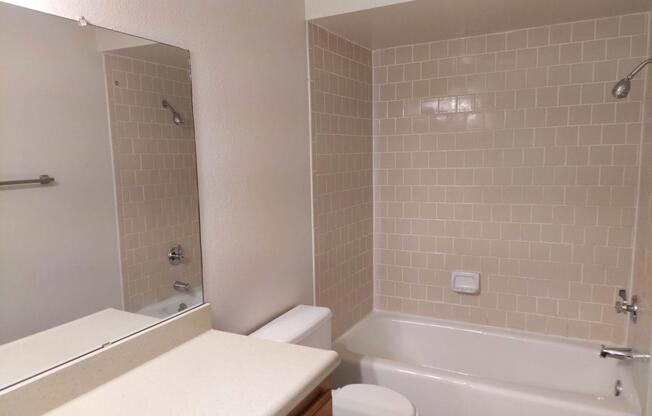 A clean, well-lit bathroom featuring a bathtub with a shower, a large mirror above a countertop, and beige tiled walls. The space includes a toilet and a towel rack. The colors are neutral, giving the room a fresh and inviting feel.