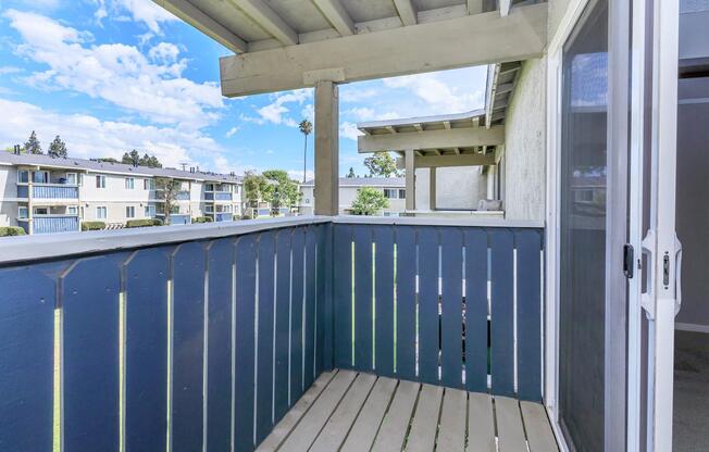 A balcony with wooden flooring and blue-painted railings, overlooking an apartment complex with multiple buildings. The sky is bright with scattered clouds, and there are trees and shrubs visible in the background. The view captures a sense of openness and tranquility.