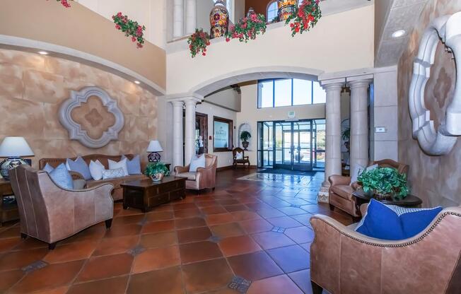 An elegant lobby featuring arched doorways, decorative columns, and large windows. The space is furnished with comfortable seating, including leather sofas and stylish armchairs. Potted plants and floral decorations add a touch of color, while warm tile flooring enhances the inviting atmosphere.