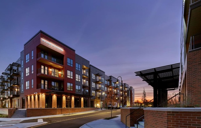 A corner street view of River Station's apartment buildings at sunset with trees and the capitol building in the background.