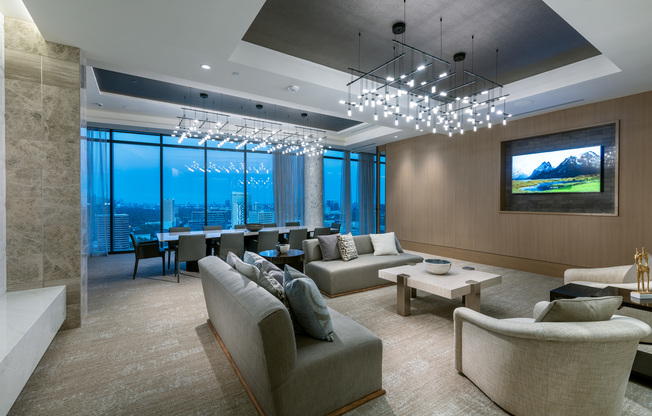Lounge area with a 12-person dining table, a seating area with two couches, and two armchairs, a large HDTV mounted on the wall, high ceilings, and a full-width floor-to-ceiling window with a city skyline view.