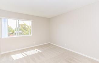 A bright, empty room with light beige walls and carpet. A large window lets in natural light, illuminating the space. Simple and clean design with no furniture, creating a minimalist atmosphere.