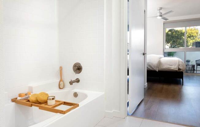 A white bathroom with a tub and a bedroom in the background. at Elements Apartments*, Irvine, CA