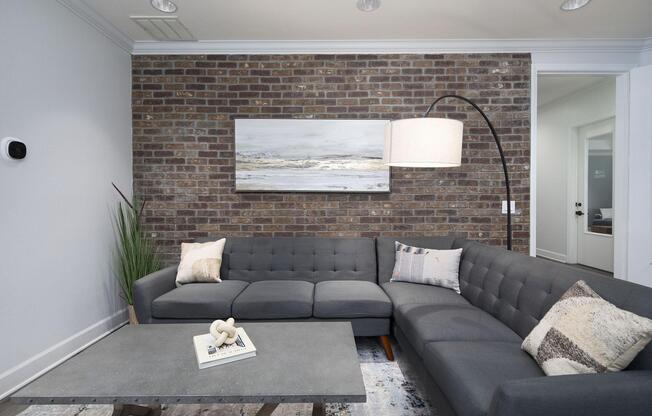 A modern living room featuring a gray sectional sofa with accent pillows, a minimalist coffee table, and a large artwork of a serene landscape on a brick wall. A stylish floor lamp curves over the sofa, and there is a decorative plant in the corner. The overall decor is contemporary and inviting.