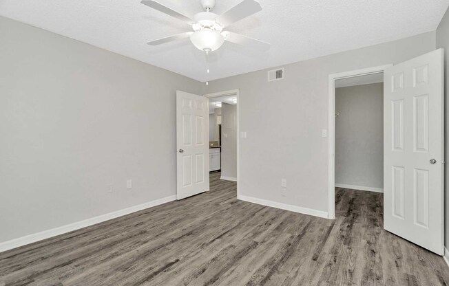 An interior view of a room featuring a ceiling fan, light-colored walls, and laminate flooring. There are two open doors leading to different areas, with one door opening to a visible space beyond. The overall design is modern and minimalistic, providing a bright and airy atmosphere.