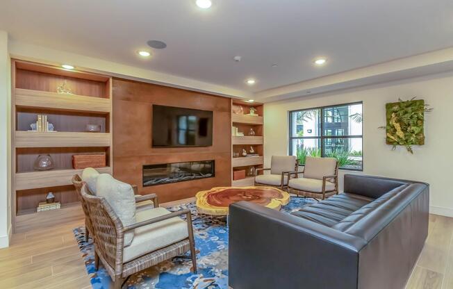 A modern living room featuring a large flat-screen TV mounted on a wooden wall unit. There are two stylish armchairs and a sleek black sofa, arranged around a unique wooden coffee table. The room is decorated with plants and has a vibrant area rug, providing a cozy and inviting atmosphere. Natural light filters through large windows.