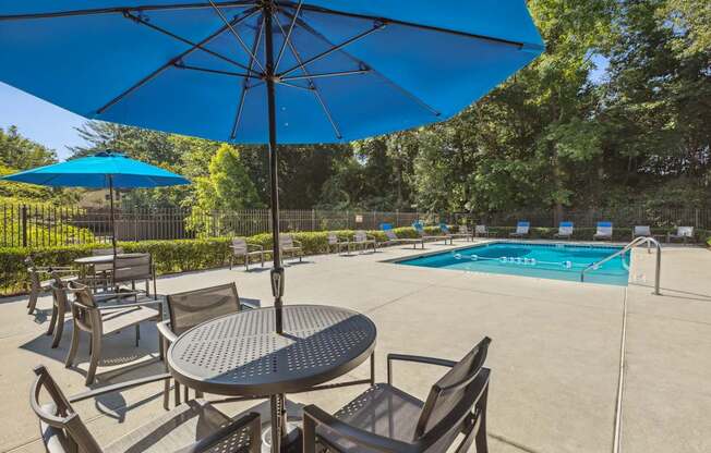 A blue umbrella shades a table and chairs by a pool.