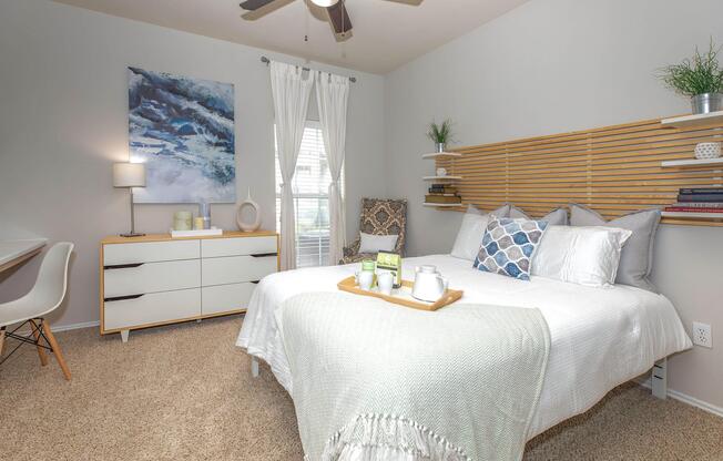 A cozy bedroom featuring a queen-size bed with decorative pillows and a soft throw. A minimalist nightstand and dresser in a light wood finish are present, along with a white desk and chair. The room has soft lighting, a fan, and wall art, creating a welcoming and stylish atmosphere.