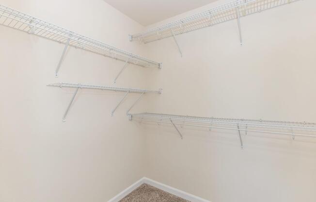 Empty closet with white wire shelving mounted on beige walls. The floor is covered with light-colored carpeting, and there are no items or decorations visible.