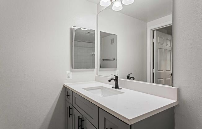 A modern bathroom featuring a white sink on a sleek gray countertop. The wall has a large rectangular mirror above the sink, with three light fixtures above it. A white door is partially visible in the background, and the walls are painted in a light gray color, creating a clean and contemporary atmosphere.