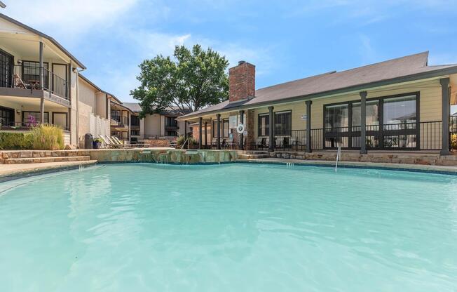 A serene swimming pool surrounded by landscaped areas, with a stone pathway leading to a charming building. The pool has a gentle waterfall feature and is bordered by a few trees. Adjacent buildings feature balconies, enhancing the inviting atmosphere of the outdoor space. Bright blue sky overhead adds to the tranquil setting.