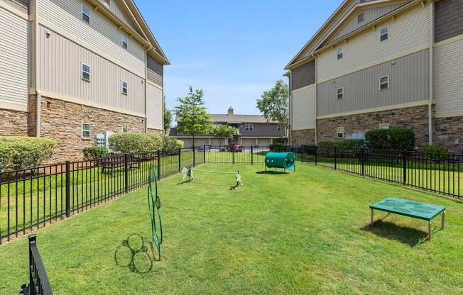 A black fence surrounds a green lawn with a tennis court and a bench.