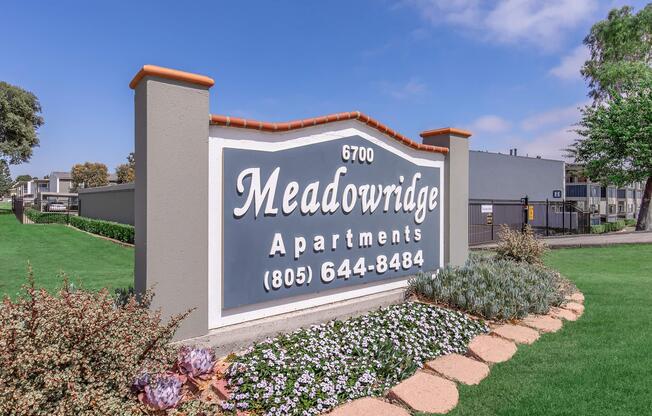 Sign for Meadowridge Apartments featuring the address 6700, phone number (805) 644-8484, surrounded by landscaped grass and flower beds. The background includes blue skies and trees, adding to the welcoming atmosphere of the apartment complex.