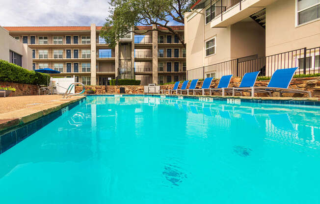 A pool area with lounge chairs, a patio table and chairs with a blue umbrella and stainless steel BBQ in front of apartment buildings at Princeton Court Apartments in the Vickery Midtown neighborhood of Dallas, TX.