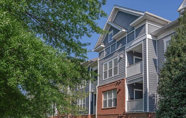 A modern, multi-story apartment building featuring a combination of gray siding and brick accents. The building has white balconies and is surrounded by trees and greenery under a clear blue sky.