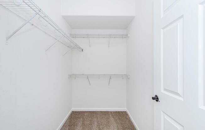 Empty closet with white walls and a carpeted floor. There are two wire shelving units mounted on the walls, and a closed door with a black doorknob is visible on the right. The space appears clean and well-lit, suitable for storage.