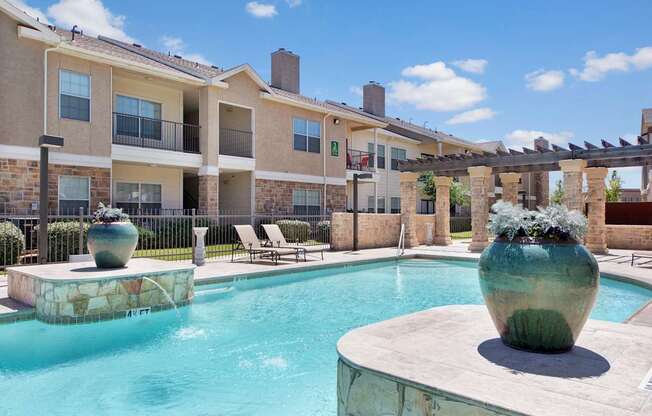 A resort-style outdoor pool area here at Mission Green showcasing clear blue water, a spacious stone pool deck, large decorative planters, shaded pergola columns, lounge chairs, and surrounding apartment buildings under a bright sky, creating a relaxing community space for residents to unwind.