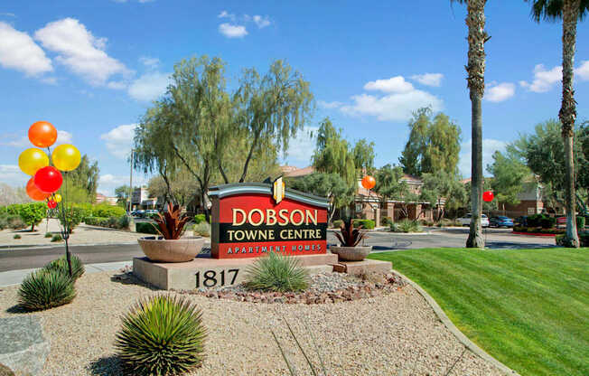 A welcoming community entrance here at Dobson Towne Center featuring a bold monument sign with Apartment Homes lettering and the address 1817, surrounded by desert landscaping, decorative rock beds, sculpted shrubs, vibrant balloons, manicured green lawn, and tall palm trees set against a bright blue Arizona sky.