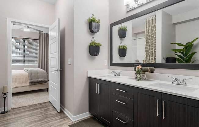 A bathroom with a large mirror, a sink, and a bedroom with a bed and window.