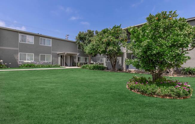A well-maintained outdoor area featuring a vibrant green lawn, surrounded by landscaped flower beds. Two gray apartment buildings are visible in the background, with trees providing shade and adding to the peaceful ambiance of the space. The sky is clear with some clouds.