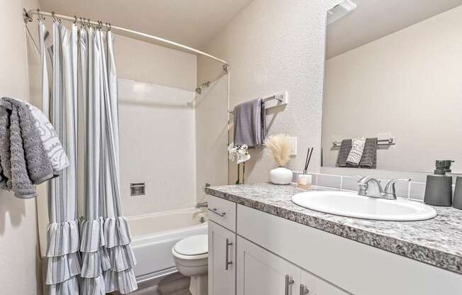 A bathroom with a white sink, toilet, and a grey shower curtain A row of apartment buildings with green lawns in front A bedroom with a bed, nightstand, and window at Wildflower, Kennewick, WA 99336.