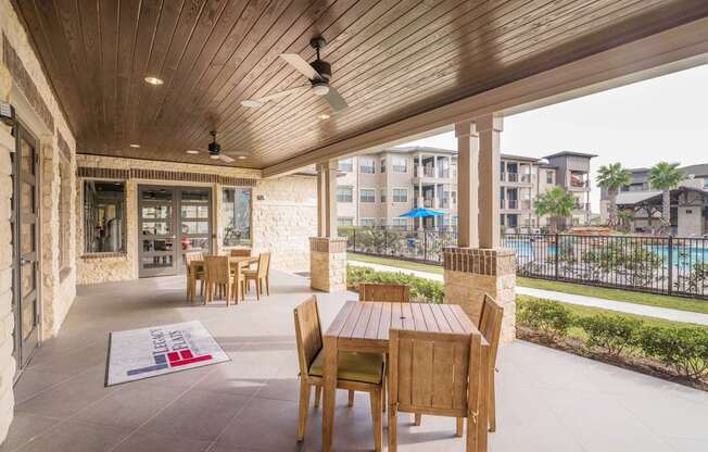 A wooden table and chairs are on a patio with a pool and buildings in the background.