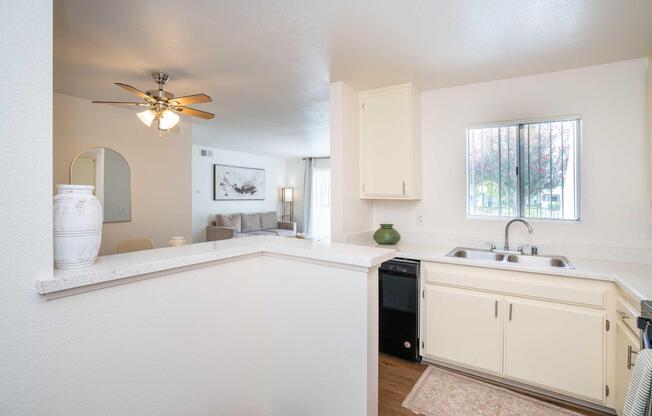 A well-lit kitchen featuring white cabinets, a double sink, and a window with blinds. A beige rug is placed in front of the sink. In the background, a living area is partially visible with a beige couch and decorative wall art. A ceiling fan is installed in the kitchen area.